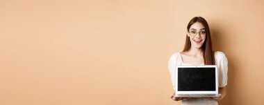 Gözlüklü, dizüstü bilgisayarlı, gülümseyen, bej arka planda duran mutlu bir kadın..