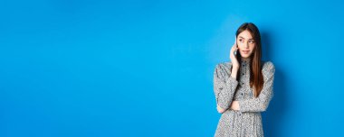 Modern, güzel bir kadın cep telefonuyla konuşuyor, logoya bakıyor ve telefon ediyor, mavi arka planda duruyor..