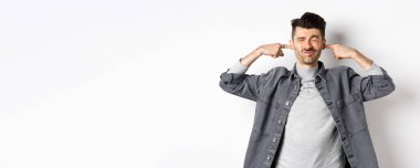 Sinirli genç adam parmaklarıyla kulaklarını tıkar ve gürültüden yüzünü buruşturur, gürültücü komşular tarafından rahatsız edilir, beyaz arka planda gürültülü bir yerde durur..