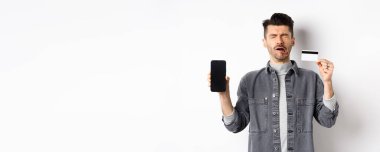 Üzgün ağlayan adam boş cep telefonu ekranı ve plastik kredi kartı gösteriyor, beyaz arka planda perişan duruyor..