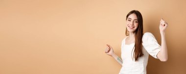 Mutlu kadın dans ediyor ve eğleniyor, kapalı gözler ve gülümsüyor, bej arka planda duruyor..