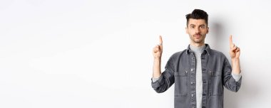 Bıyıklı, ciddi bir beyaz adam. Parmaklarını boş yere doğrultuyor ve beyaz arka planda kameraya bakıyor..