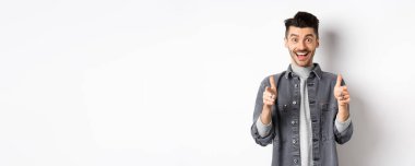 Heyecanlı genç adam başparmağını kaldırıyor ve mutlu gülümsüyor, övgüler yağdırıyor, seni motive ediyor, havalı ürünler öneriyor, beyaz arka planda duruyor..