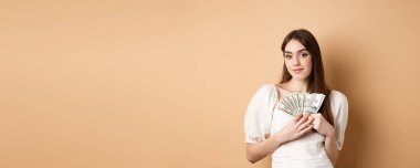 Gülümseyen zengin kadın dolarları gösteriyor ve kameraya mutlu bakıyor, para kazanıyor ya da hızlı kredi veriyor, bej arka planda duruyor..