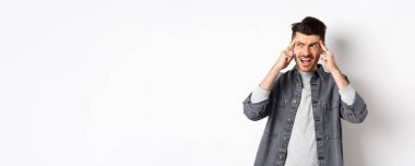 Sıkıntılı genç adam kafaya dokunuyor ve bağırıyor, hayal kırıklığına uğramış görünüyor, beyaz arka planda duruyor..