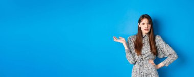 Kafası karışmış ve sinirli kız tartışıyor, elini kaldırıyor ve kaşlarını çatıyor. Mavi arka planda dikilmenin ne önemi var anlamıyorum..