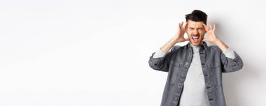 Sinirli genç adam bağırıyor ve kafaya dokunuyor, ayakta duruyor gürültüden ya da baş ağrısından rahatsız oluyor, beyaz arka planda duruyor..