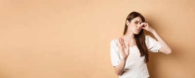 Tatminsiz kadın burnunu kötü kokudan tıkar, jesti durdurur ve tiksinti yüzünden kaşlarını çatar. Burun deliklerini nefretle kapat, bej arka planda dur.