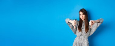 Olumsuz duygu. Uzun saçlı ve elbiseli, hayal kırıklığına uğramış bir kadın, baş parmaklarını aşağı gösteriyor ve suratını ekşitiyor, hoşnutsuzluğunu ifade ediyor, mavi arka planda duruyor..