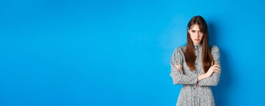 Elbiseli kızgın genç bir kadın adaletsiz hissediyor, kollarını göğsünde kavuşturuyor ve küskün, mavi arka planda duruyor..