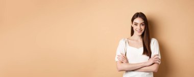 Göğsünde çapraz kollar olan, gülümseyen samimi kız, profesyonel gibi görünüyor, bej arka planda kendine güveniyor..