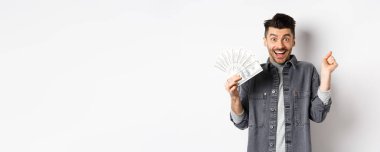 Neşeli genç adam heyecandan zıplıyor ve dolarları gösteriyor, ödül parası kazanıyor, para kazanıyor ve seviniyor, beyaz arka planda duruyor..