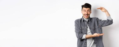 Heyecanlı adam logoya bakıyor ve büyük bir şey gösteriyor, büyük bir nesne şekillendiriyor ve şaşkın bir şekilde gülümsüyor, beyaz arka planda kot ceketle duruyor..