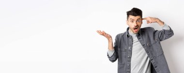 Delirdin mi sen? Sinirli bir adam aptalca davranışları için insanları azarlıyor, baş tapınağını işaret ediyor ve kafası karışmış bir şekilde elini kaldırıyor, beyaz arka planda dururken şikayet ediyor..