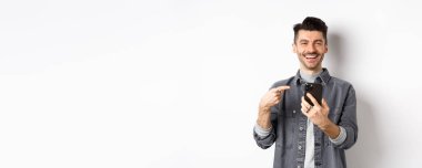 Bıyıklı yakışıklı beyaz adam akıllı telefonu işaret ediyor, gülümsüyor ve cep telefonuyla iyi bir anlaşma yapıyor, beyaz arka plana karşı mutlu duruyor..