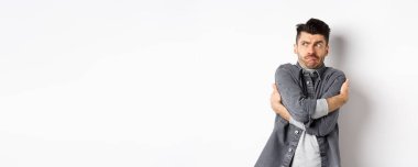 Memnun olmayan genç adam kendini rahatsız hissediyor, kendine sarılıyor ve utanç verici bir şeye bakıyor ya da teşhir ediyor, beyaz arka planda rahatsız bir şekilde duruyor..