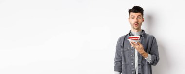 Yakışıklı doğum günü çocuğu pastaya mum üflüyor ve dilek tutuyor, kameraya bakıp, beyaz arka planda duruyor..