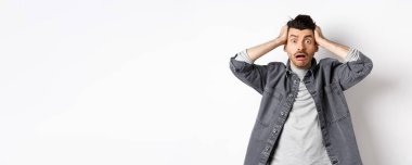 Telaşlı genç adam kameraya bakıyor, endişeli ve sıkıntılı görünüyor, büyük bir problemi var, beyaz arka planda panikliyor..