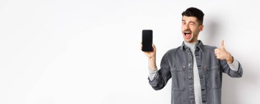 Yakışıklı adam göz kırpıyor ve boş cep telefonu ekranını başparmağıyla gösteriyor. Uygulama veya çevrimiçi anlaşma öneriyor, beyaz arkaplana karşı duruyor..