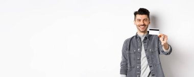 Yakışıklı adam plastik kredi kartı gösteriyor ve gülümsüyor, tatmin olmuş müşteri beyaz arka planda duruyor..