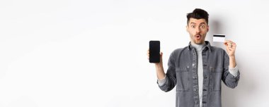 Plastik kredi kartı ve cep telefonu ekranıyla beyaz arka planda duran heyecanlı yakışıklı adam..