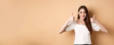 - Çok iyi. Mutlu ve tatmin olmuş genç bir kadın, olumlu gülümsüyor, onaylıyor ve mükemmel bir seçim yapıyor, seni övüyor, iyi bir jest yapıyor, bej arka plan yapıyor..