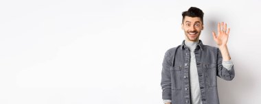 Dost canlısı stil sahibi adam merhaba diyor ve elini sallıyor, neşeli bir şekilde gülümsüyor, selam veriyor, beyaz arka planda duruyor..