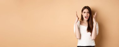 Meyveli kadın bağırıyor, el sıkışıyor, kızgın görünüyor, kızgın görünüyor, bej arka planda sıkıntılı duruyor..