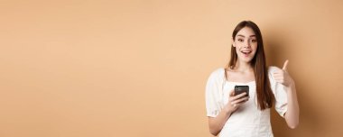 İnternetten alışveriş konsepti. Mutlu genç bir kadın baş parmağını gösteriyor ve cep telefonu kullanıyor, gülümsüyor lütfen, bej arka planda duruyor..