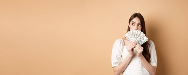 Dolarları tutan ve boş alana bakan düşünceli bir kız. Parayla ne alacağını düşünüyor, alışverişi planlıyor, bej arka planda duruyor..