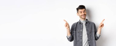 Bıyıklı genç yakışıklı adam iki logo gösteriyor, kameraya mutlu bir şekilde gülümsüyor, beyaz arka planda günlük giysiler içinde duruyor..