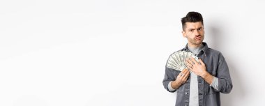 Açgözlü adam kaşlarını çatıyor ve dolarları kucaklıyor, parayı paylaşmak istemiyor, beyaz arka planda duruyor..