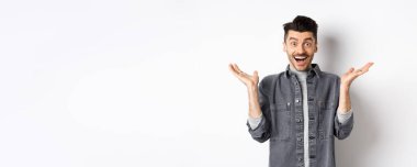 Heyecanlı genç adam sevinçten havalara uçuyor, ellerini yana açıyor ve mutlu görünüyor, şaşkın şaşkın gülüyor, beyaz arka planda duruyor..