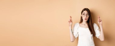 Heyecanlı kız umut rüyası gerçekleşiyor, parmaklarını çapraz yapıp dua ediyor, bej arka planda duruyor..