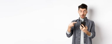 Akıllı telefondan havalı bir şey gösteren genç adam etkilenmiş bir şekilde telefona bakıyor ve beyaz arka planda duruyor..