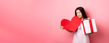 Sevgililer Günü konsepti. Romantik Asyalı genç kız beyaz bir günde sevgilisinden hediyeler alır. Büyük kırmızı kalp kartına sarılır ve kutuda sunulur. Güler yüzlü, pembe arka plan..
