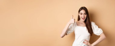 Şaşırmış genç bir kadın ortaya çıkıyor, evet diyor ve harika bir ürünü övüyor, onaylıyor, bej arka planda tatmin olmuş bir şekilde duruyor..