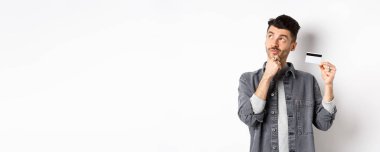 Sol üst köşedeki logoya bakıp elinde plastik kredi kartı tutan, alışveriş yapmayı düşünen, beyaz arka planda duran düşünceli bir adam..