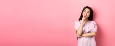 Dalgın gülümseyen Asyalı kız seçim yapıyor, sol üst köşedeki logoya bakıyor ve düşünüyor, bir fikri var, pembe arka planda kurnazca duruyor..