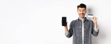 Plastik kredi kartı ve cep telefonu ekranıyla beyaz arka planda duran heyecanlı yakışıklı adam..