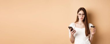 Kafası karışmış gözlüklü bir kız cep telefonu okuduktan sonra kameraya kaşlarını çatıyor, elinde kahve fincanı tutuyor, bej arka planda hoşnutsuz duruyor..