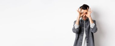 Komik adam el dürbününden bakıyor, muhteşem tanıtımlara bakıyor, hayret ediyor ve kameraya bakıyor, heyecanlı, beyaz arka planda duruyor..