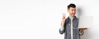 Dizüstü bilgisayardaki komik genç adam görüntülü sohbet. Bilgisayar kamerasına bakıp gülümsüyor. Beyaz arka planda duruyor..