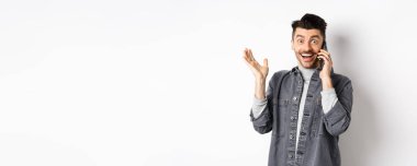 Heyecanlı adam cep telefonuyla konuşuyor ve iyi haberler duyuyor, seviniyor ve mutlu bir gülümsemeyle elini kaldırıyor, beyaz arka plana karşı duruyor..
