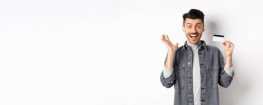 Heyecanlı adam sevinçten çığlık atıyor ve plastik kredi kartı gösteriyor, alışveriş yaparken seviniyor, beyaz arka planda duruyor..