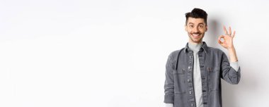 Gülümseyen yakışıklı adam iyi ürün gibi tamam işareti gösteriyor, reklam öneriyor, beyaz arka planda tatmin olmuş duruyor..
