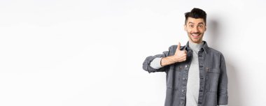 Heyecanlı yakışıklı adam baş parmağını gösteriyor ve gülümsüyor, iyi bir şey gibi, harika seçimi övüyor, tatmin olmuş ve mutlu görünüyor, beyaz arka planda duruyor..