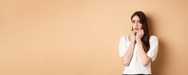 Korkmuş ürkek kız korkmuş görünüyor, korkudan titriyor ve kameraya bakıyor, kameraya korkmuş görünüyor, bej arka planda giyinmiş duruyor..