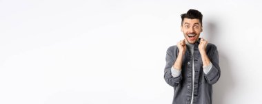 Heyecanlı genç adam mutluluktan atlıyor, şaşırmış bir şekilde gülümsüyor ve el ele tutuşuyor, beyaz arka planda duruyor..