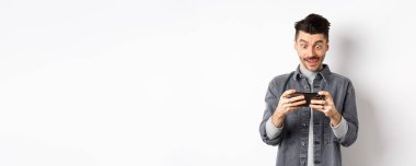 Heyecanlı adam cep telefonuyla video oyunu oynuyor odaklanmış yüzü, akıllı telefon ekranını dinliyor, cep telefonunu yatay olarak tutuyor, beyaz arka plan.